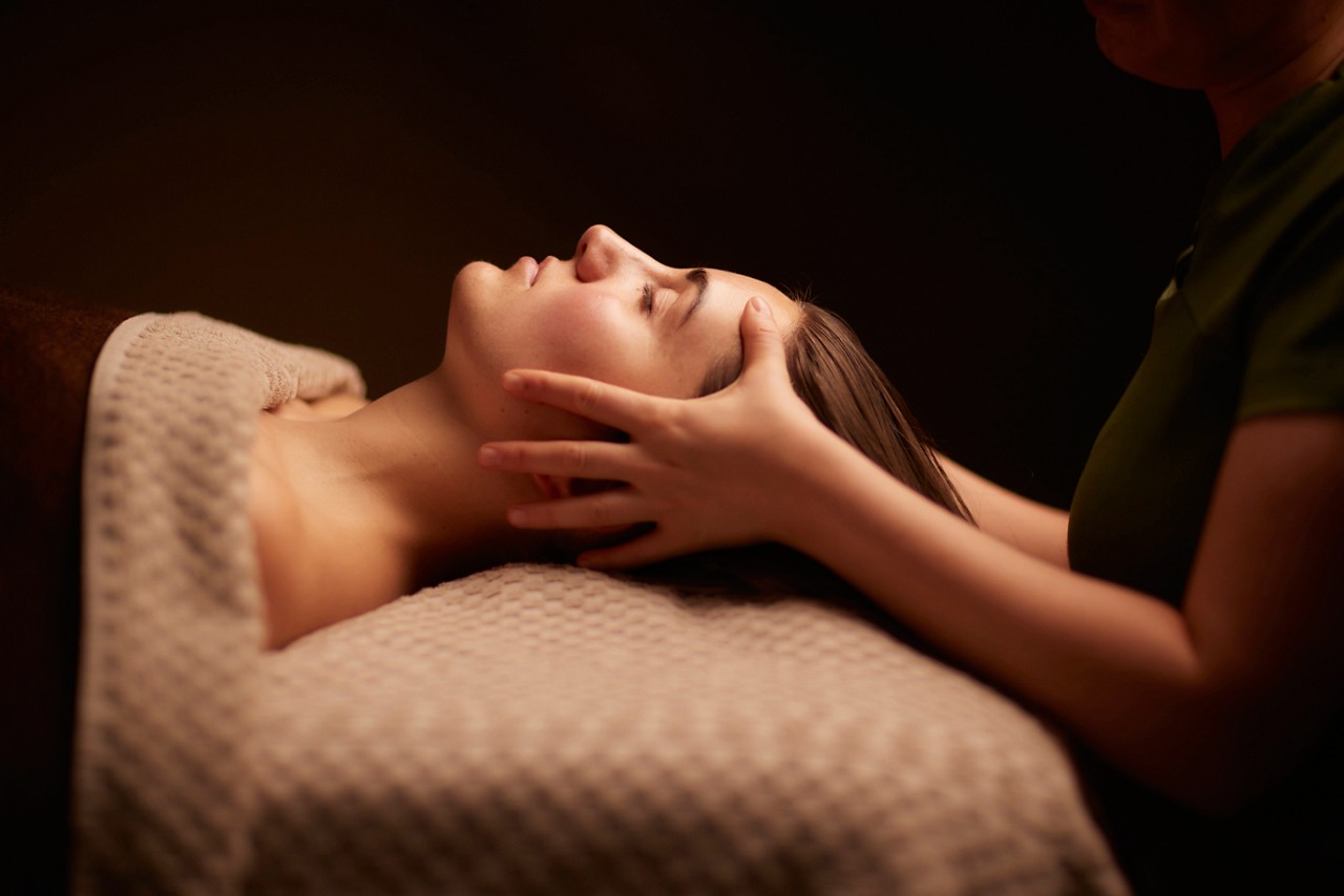 A person’s face, eyes closed, receives a gentle head massage; a therapist’s hands cradle the jaw and scalp while they lie under a textured blanket in a dim spa setting.